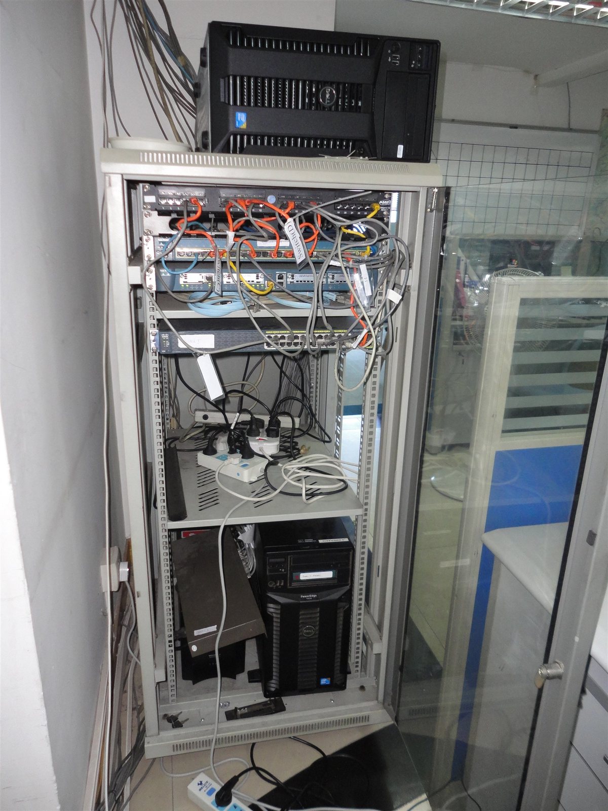 Floor-standing network cabinet in an office server corner before relocation, with tower servers and power strips